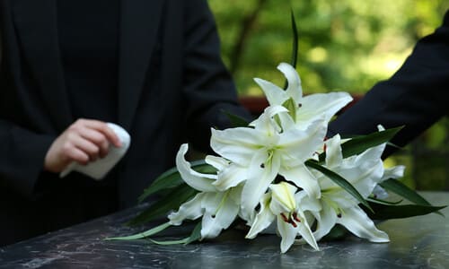 White flowers on top of a car accident victim's casket, with two mourning loved ones in the background.