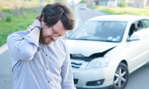 A driver standing outside his car after an accident, holding the back of his neck in pain.