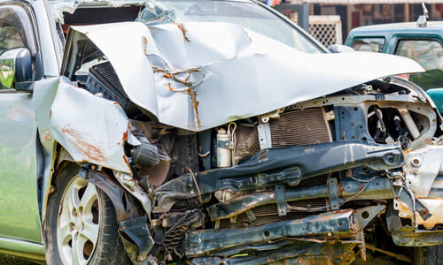 A white car with a crumpled front and totaled engine block sitting on the side of the road after an accident.