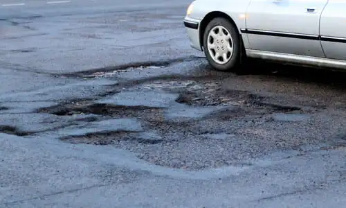 A grey car driving past a damaged section of road on a rainy afternoon.