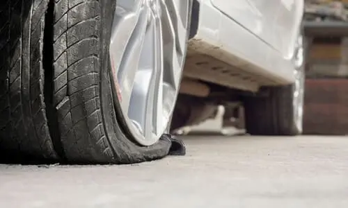 A closeup of a grey sedan with a damaged tire in front of a garage.