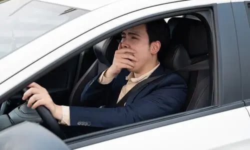 A fatigued driver yawning while behind the wheel of his silver sedan.