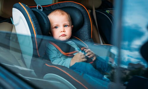 A baby looking out a car window while seated in the car seat.
