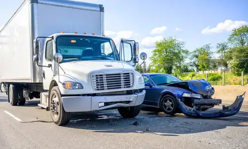 A dark blue family sedan having lost its front bumper in a highway accident with a large white semi truck.