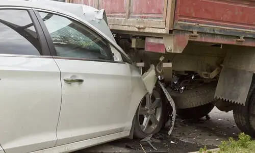 A grey car having sustained severe damage after crashing into the rear of a truck at high speed.