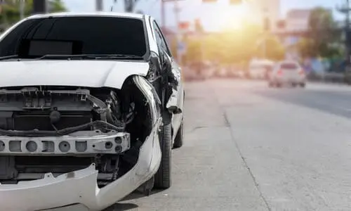 A white car in a city street with its front bumper missing after a speeding racer collided with its corner.