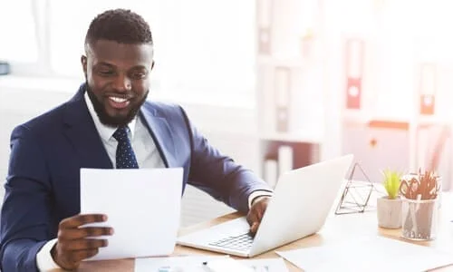 A personal injury attorney smiling as he holds up a client's insurance policy while working on a laptop.